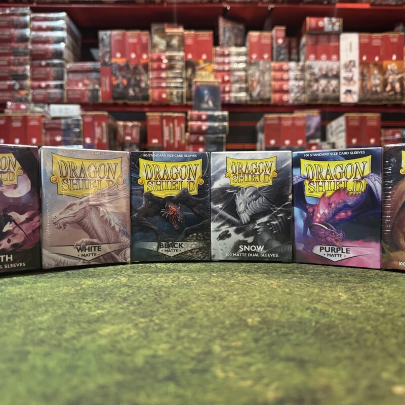 Six boxes of Dragon Shield card sleeves on a gaming table: Wraith, White, Black, Snow, Purple and Peach.