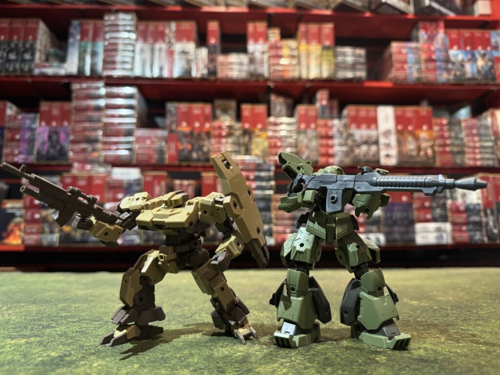 Tan and green 30 Minutes Missions robot models posed with rifles on a gaming table in front of hobby shop shelves.
