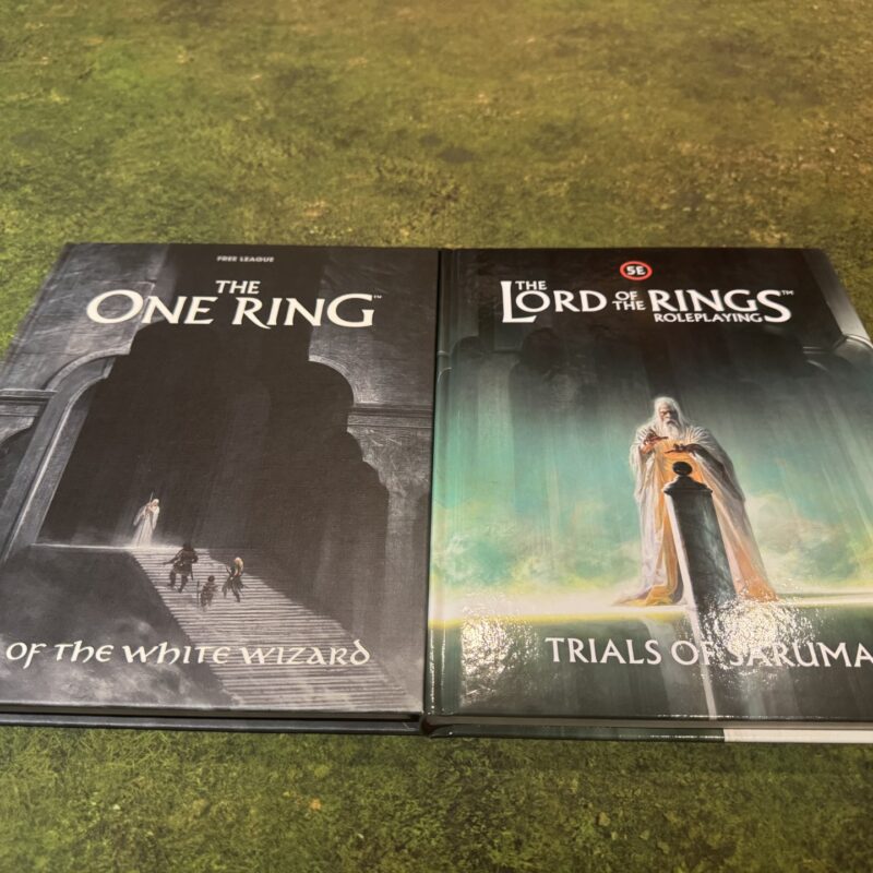 Hands of the White Wizard for The One Ring and Trials of Saruman for The Lord of the Rings Roleplaying 5E, side by side on a green gaming mat.