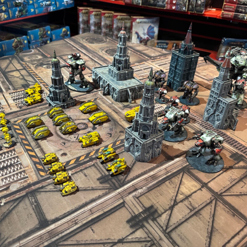 Overhead view of yellow tanks and small walkers beside larger green, purple and red walkers among grey model towers.