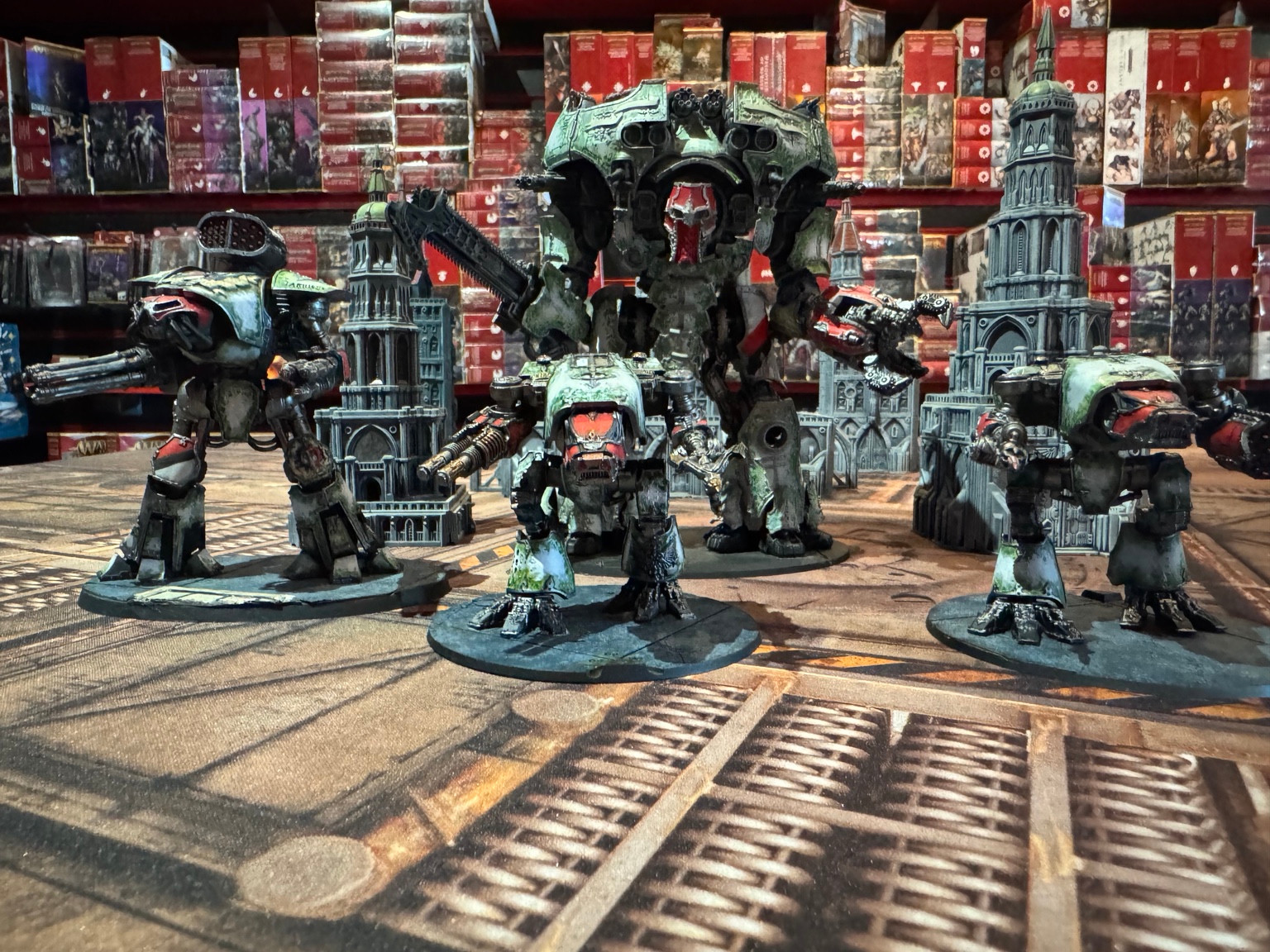 Green and red Titans of different sizes positioned among grey Gothic buildings on a Legions Imperialis tabletop.