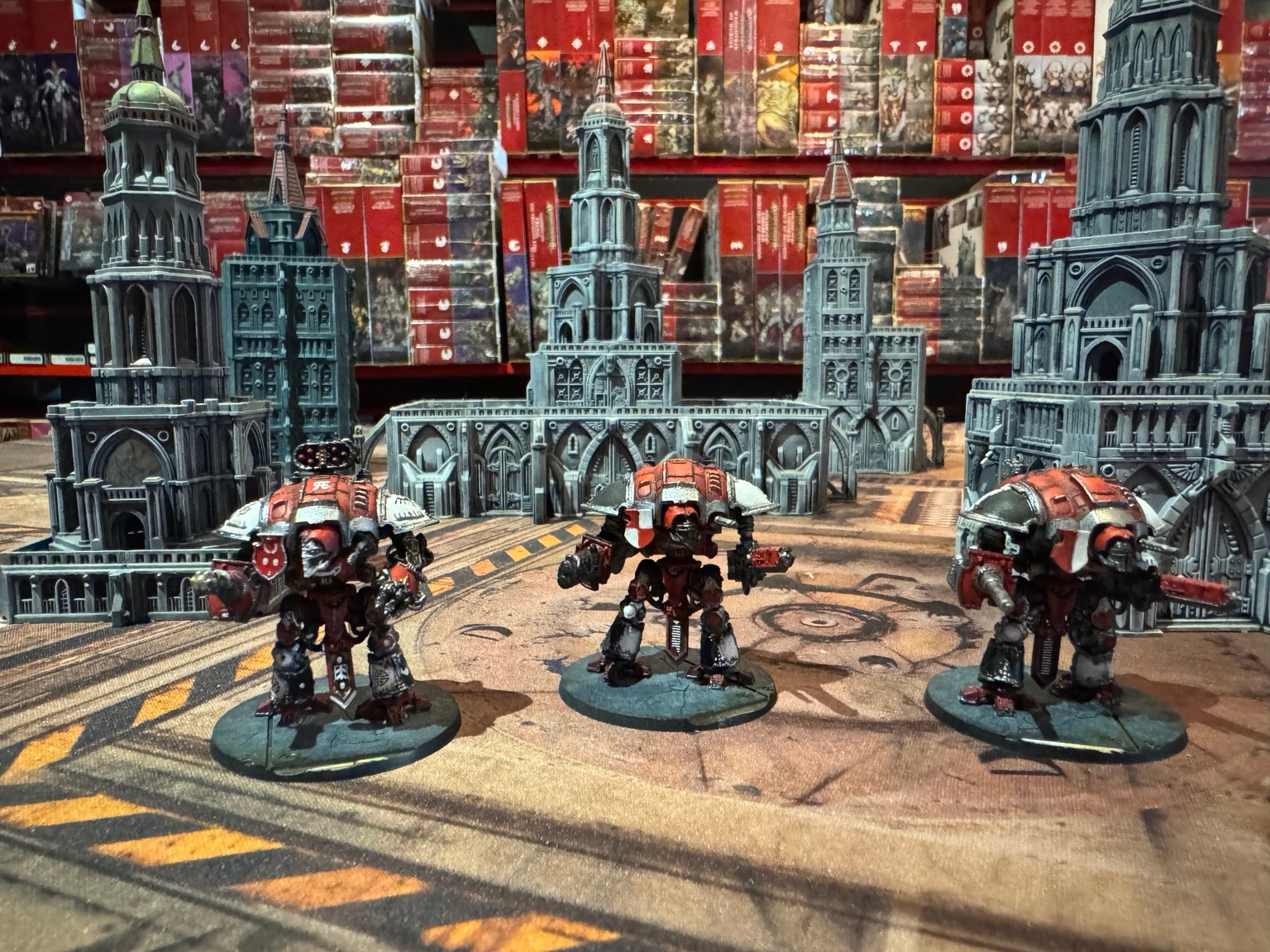 Three red and white Knights on round bases among grey Gothic buildings on a Legions Imperialis tabletop.